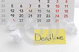 Yellow paper reminder note with text Deadline, paper balls against calendar on the white desk