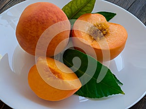 Yellow peaches full and cut on white plate with green leaves, on wooden table. Closeup, fruits