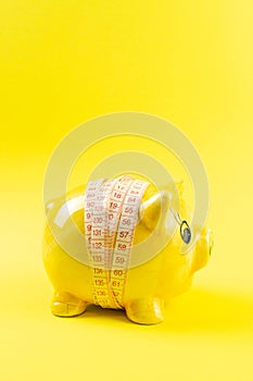 Yellow piggy bank in the form of a pig wrapped with measuring tape on a yellow background. The concept of saving money for the