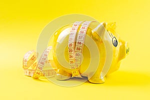 Yellow piggy bank in the form of a pig wrapped with measuring tape on a yellow background. The concept of saving money for the