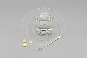 Yellow pills, thermometer and glass of water on gray background