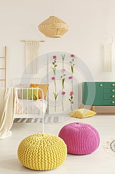 Yellow and pink pouf in the middle of boho female bedroom with green cabinet, flower board, single bed and macrame on the wall