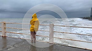 in the Yellow raincoat to watch the storm. A woman stands on the pier and looks at the big waves near the shore. The