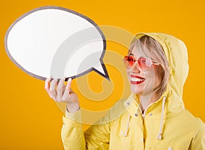 Yellow raincoat woman holding speech bubble. Bright emotions and yellow background. Pink heart shaped sunglasses