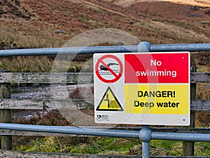 A yellow and red United Utilities sign on a steel barrier prohibits swimming in a pool above a weir on the Whitendale River