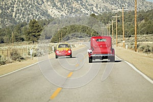 A yellow and red VW hotrod drives in opposite direction of a restored bright Red Roadster hotrod pickup truck along rural highway