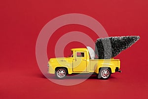 Yellow retro toy pickup carrying a Christmas tree on red background. Christmas and New Year celebration concept. Copy space,