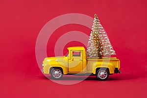 Yellow retro toy pickup carrying a Christmas tree on red background. Christmas and New Year celebration concept. Copy space,