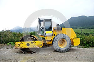 The yellow road roller with a closed cab stands on an uneven road on stones and gravel, side view.
