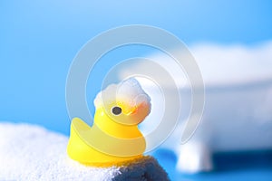 Yellow rubber duck for bathing with foam on a towel on a blue background with a bath, close up
