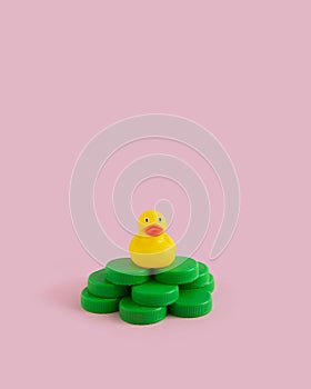 Yellow rubber duck on a green plastic waste pollution island on a pink pastel background. Polluted environments.