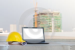 The yellow safety helmet put on the blueprint with laptop has white screen isolated at construction site with crane background
