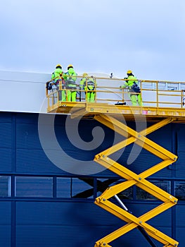 Yellow self propelled scissor lift with workers against a blue sandwich panel wall background