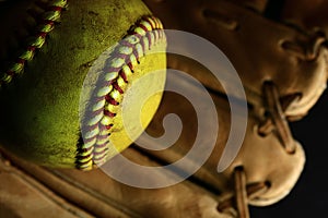 Yellow softball closeup with red seams on a brown leather glove.