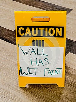 Yellow standing Caution sign sitting on parquet wood floor with white paper hand lettered Wall Has Wet Paint taped to it