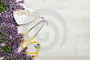 Yellow stethoscope with lilac flowers and gift box on light wooden background. Top view with copy space