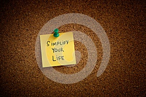 A yellow sticky note with the phrase Simplify Your Life written on it pinned to a cork board