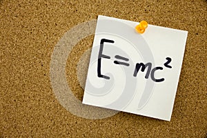 A yellow sticky note writing, caption, inscription EQUATION E EQUAL MC2 in black ext on a sticky note pinned to a cork