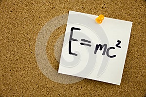 A yellow sticky note writing, caption, inscription EQUATION E EQUAL MC2 in black ext on a sticky note pinned to a cork notice boar