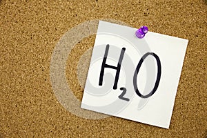 A yellow sticky note writing, caption, inscription Phrase H2O in black ext on a sticky note pinned to a cork notice board