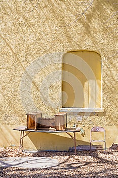 Yellow stucco vintage building has boarded window and old table with desk & chair outside.