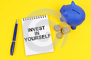 On a yellow surface, a piggy bank, coins and a notepad with the inscription - Invest in yourself