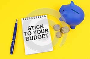 On a yellow surface, a piggy bank, coins and a notepad with the inscription - Stick to your budget