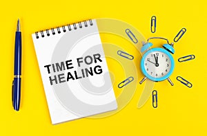 On a yellow surface there is an alarm clock, a pen and a notepad with the inscription - Time for Healing