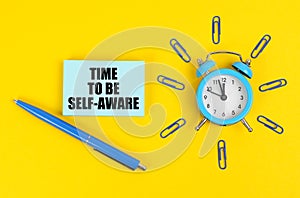 On the yellow surface there is an alarm clock, a pen and stickers with the inscription - Time to Be Self-Aware