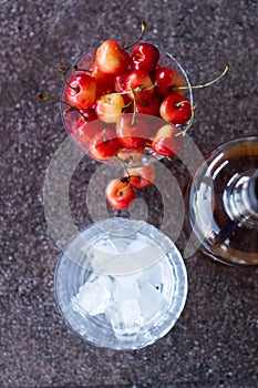 Yellow sweet cherries in a whiskey glass, ice cubes and a bottle of whiskey against a stone background. A refreshing cold cocktail