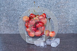 Yellow sweet cherries in a whiskey glass, ice cubes and a bottle of whiskey against a stone background. A refreshing cold cocktail