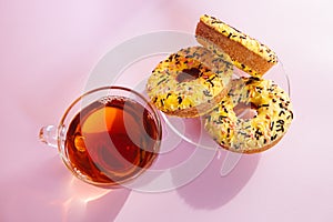 Yellow sweets - donuts biscuits and black tea in glass cup on pink table with sunshine, top view