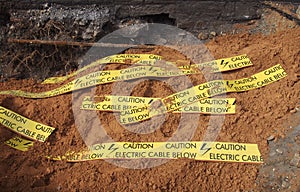 Yellow tape signs warning of buried electric cable below in a trench being excavated during roadwork and building construction