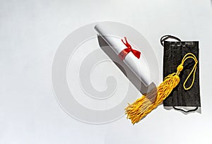 Yellow tassel from graduation cap, diploma and black protective face mask, paper scroll tied with red ribbon with bow on white