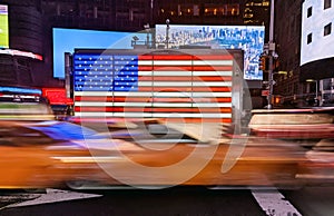 Yellow taxis driving past an American flag in Times Square, New York City with motion blur