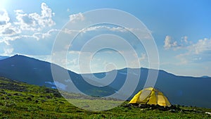 A yellow tent stands against a beautiful mountain landscape. The sun`s rays break through the clouds. Hiking in the mountains