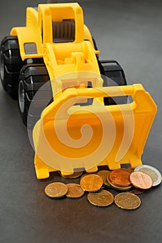 Yellow toy bulldozer, excavator carrying a lot of money - euro cent coins on dark background. Money digging, subsidies from Europe