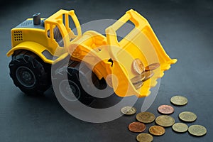 Yellow toy bulldozer, excavator carrying a lot of money - euro cent coins on dark background. Money digging, subsidies from Europe