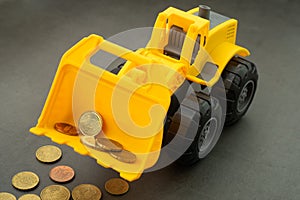 Yellow toy bulldozer, excavator carrying a lot of money - euro cent coins on dark background. Money digging, subsidies from Europe