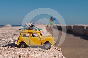 Yellow toy car with a surfboard and a shell on the breakwater. Mediterranean Sea, clear hot day.