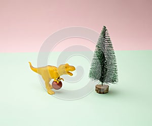 Yellow toy dinosaur tyrannosaurus rex with new year red baulbe around nack with golden rope and Christmas tree on a pink and mint