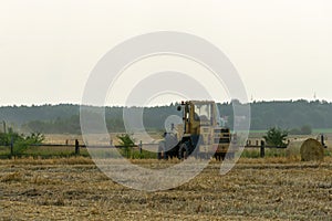 A yellow tractor with the help of a manipulator puts round bales of hay on a trailer. Transportation of hay bales in meta storage