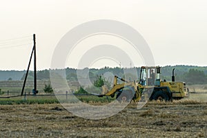 A yellow tractor with the help of a manipulator puts round bales of hay on a trailer. Transportation of hay bales in meta storage