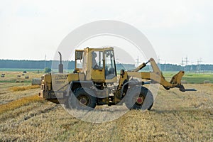 A yellow tractor with the help of a manipulator puts round bales of hay on a trailer. Transportation of hay bales in meta storage