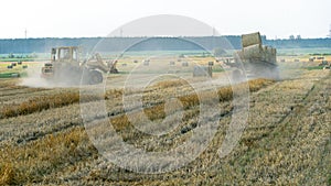 A yellow tractor with the help of a manipulator puts round bales of hay on a trailer. Transportation of hay bales in meta storage