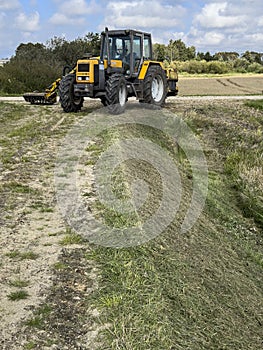 Yellow tractor mowing grass overgrowing the flood bank on the river