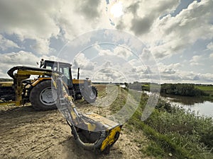 Yellow tractor mowing grass overgrowing the flood bank on the river