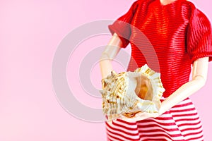 Yellow tropical seashell in woman doll hands on light pink background with copy space. Marine vacation, tourism and summer holiday