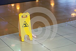 Yellow warning plate wit Caution wet floor icon on slippery tile floor st shopping mall indoors