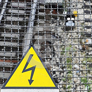 Yellow warning sign for high voltage contact wires above the rails, in the background the train station tracks. Warning icon